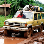**Image Prompt:** A heavily potholed, unpaved road in Guinea. A battered 4x4 vehicle is stuck in the mud, surrounded by smiling locals offering assistance. Lush green vegetation in the background. Focus on the collaborative spirit.